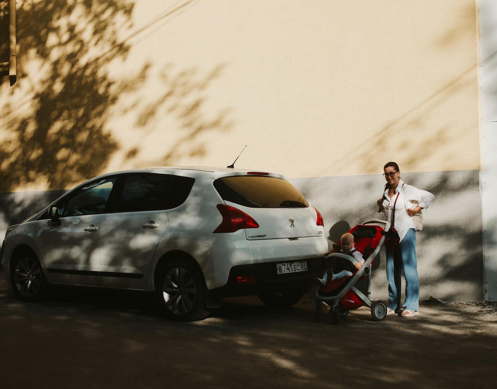 Mama z wózkiem i dzieckiem przy białym aucie na słonecznej ulicy